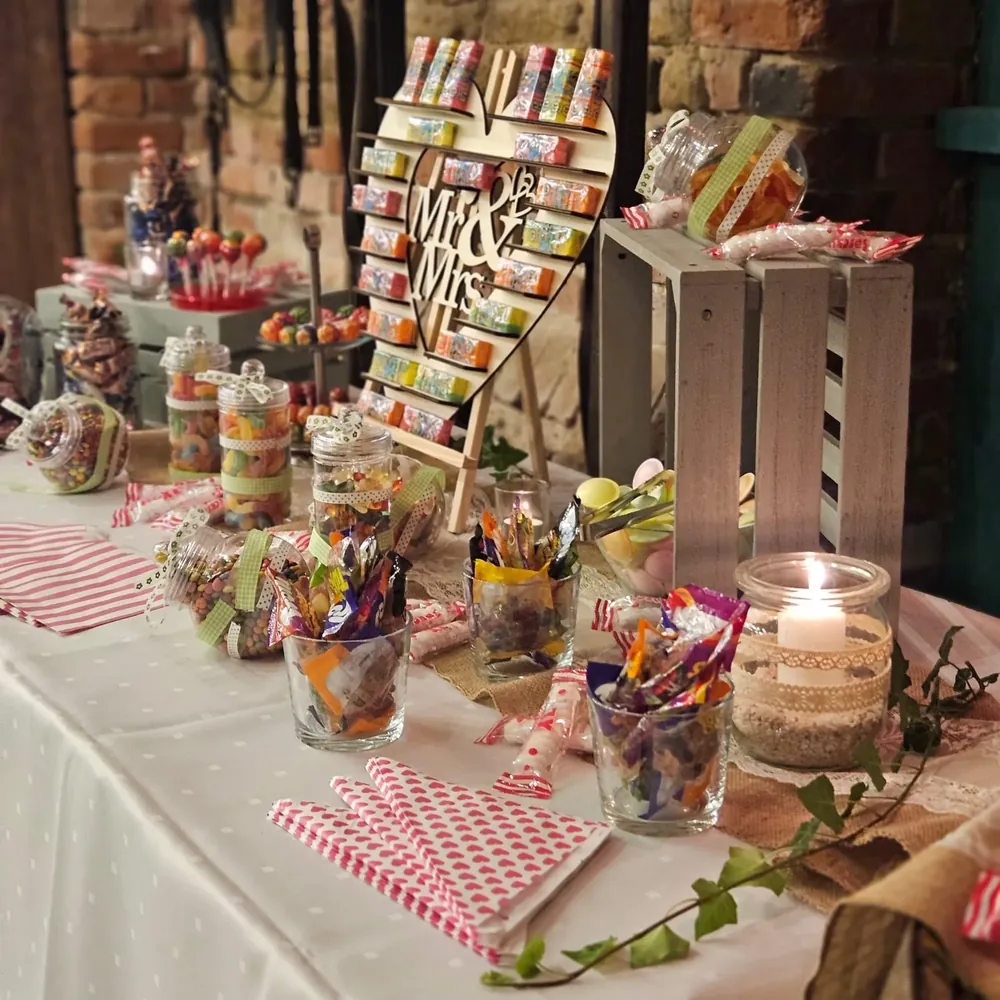 Candy Bar mit Mr-and-Mrs-Herz, Süßigkeiten in Gläsern und Kerzenlicht vor der Backsteinwand der Feierscheune