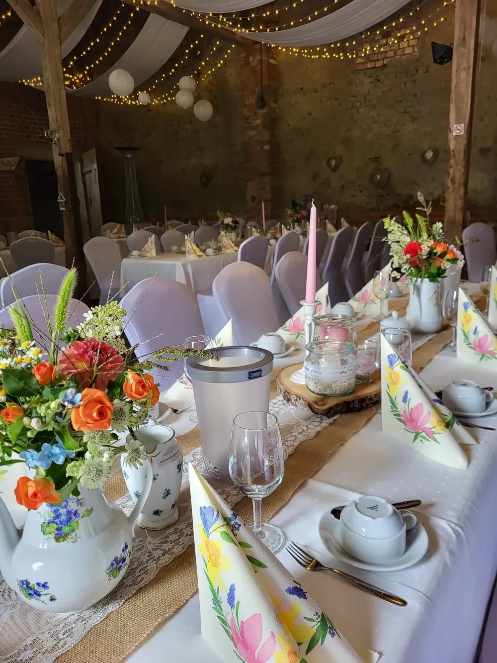 Festtafel in der Feierscheune unter Stoffbahnen und Lichterketten mit Wiesenblumen, rosa Kerze und Blumenservietten