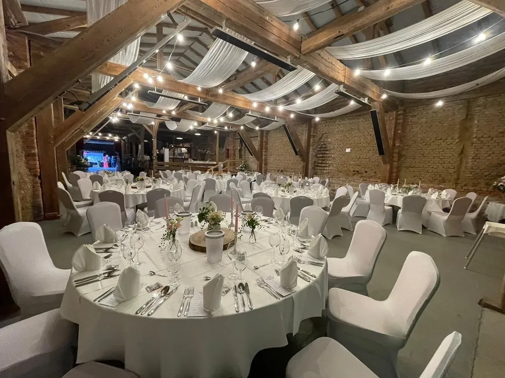 Runde Hochzeitstische mit weißen Hussen unter Holzbalken, Stoffbahnen und Lichterketten im Hauptbereich der Feierscheune