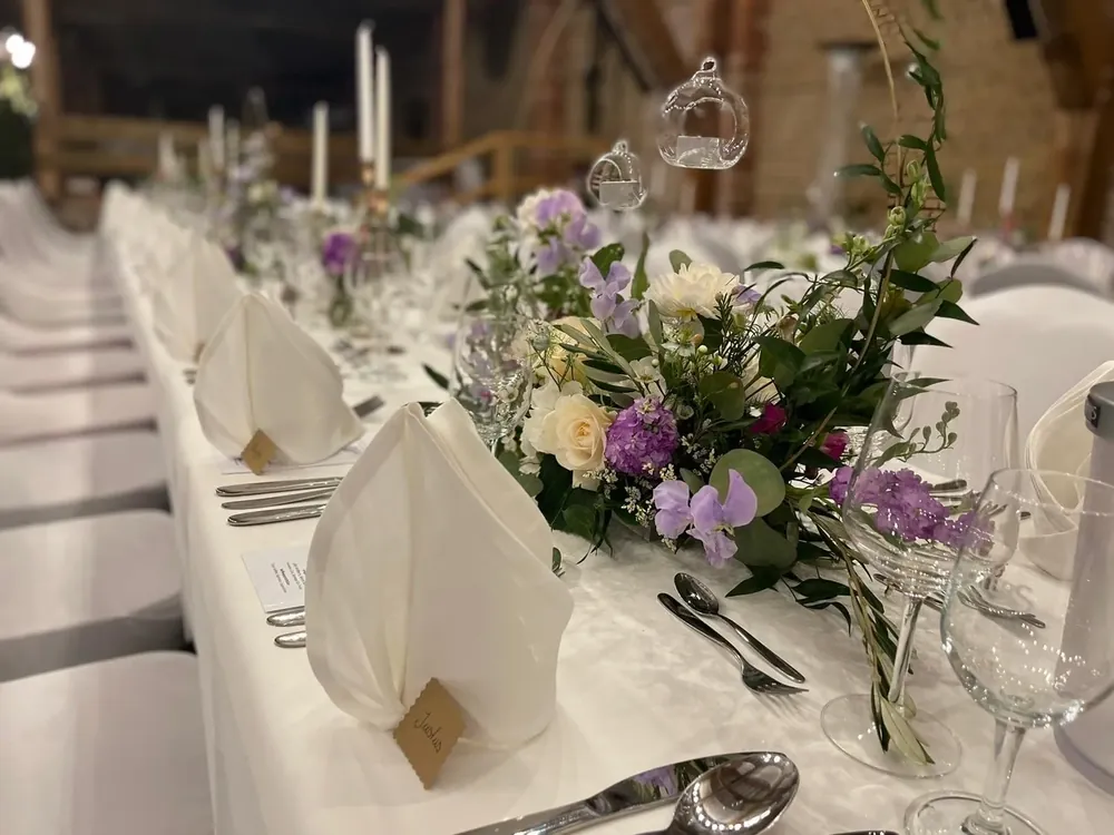 Hochzeitstafel in der Feierscheune mit Blumenschmuck in Flieder und Weiß, gefalteten Servietten und hängenden Glaswindlichtern