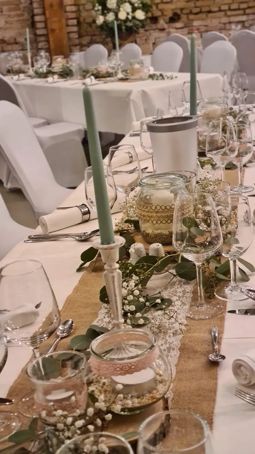 Festtafel mit grünen Stabkerzen, Eukalyptus, Schleierkraut und Windlichtern auf Juteläufer vor Backsteinwand