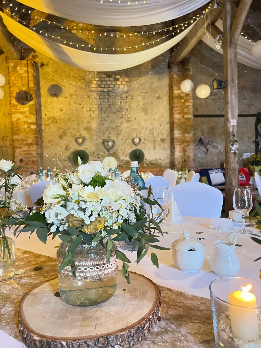Tischdekoration mit weißen Rosen im Glas mit Spitzenband auf einer Baumscheibe unter Stoffbahnen und Lichterketten in der Feierscheune