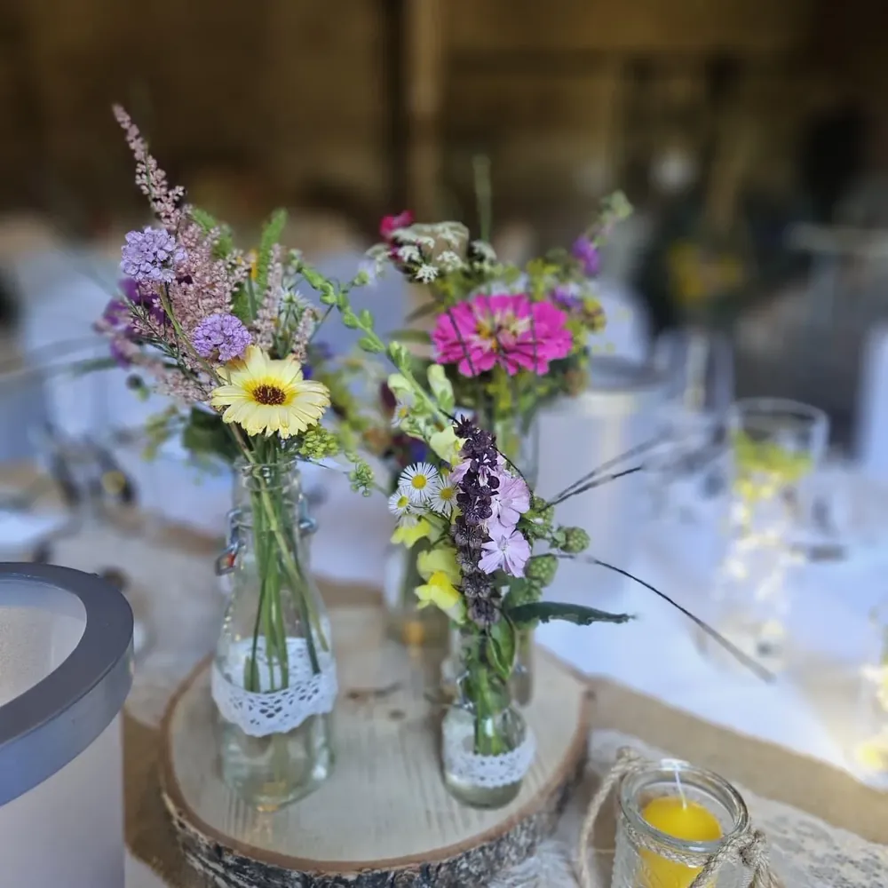 Wiesenblumen in Flaschen mit Spitzenband und Bienenwachskerze auf Baumscheibe bei einer Hochzeit in der Feierscheune