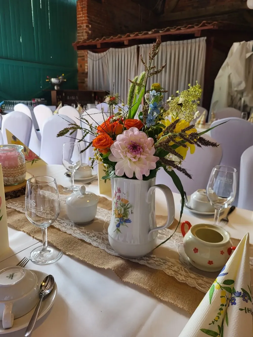 Blumenstrauß mit Dahlie und Wiesenblumen in einer Porzellankanne auf Juteläufer mit Spitze, gedeckter Tisch in der Feierscheune Arendsee